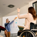 Back view of businesswoman with disability attending a seminar and raising her hand to answer a question.